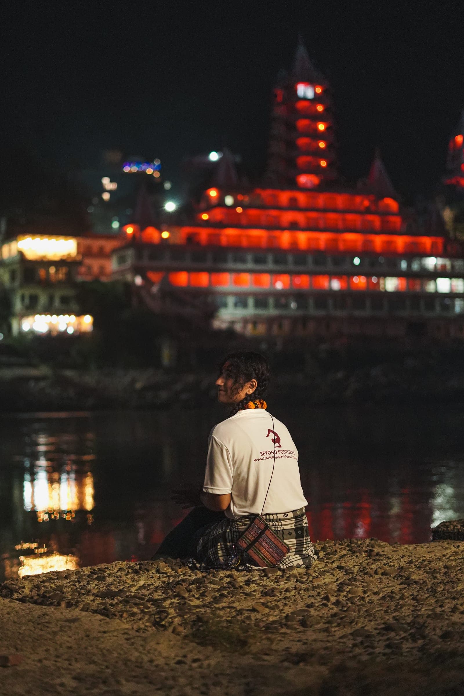 A student sitting by the Ganga at night, the lit temple across the water