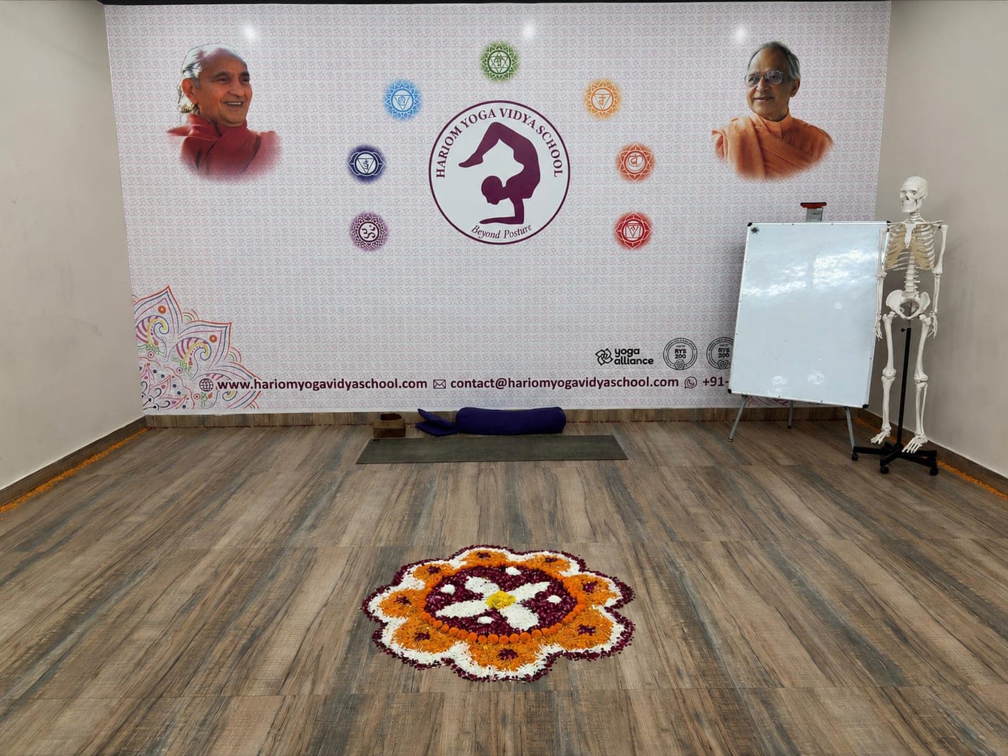 The founders' portraits and the school emblem on the hall's welcome wall, above a flower rangoli laid out on the floor