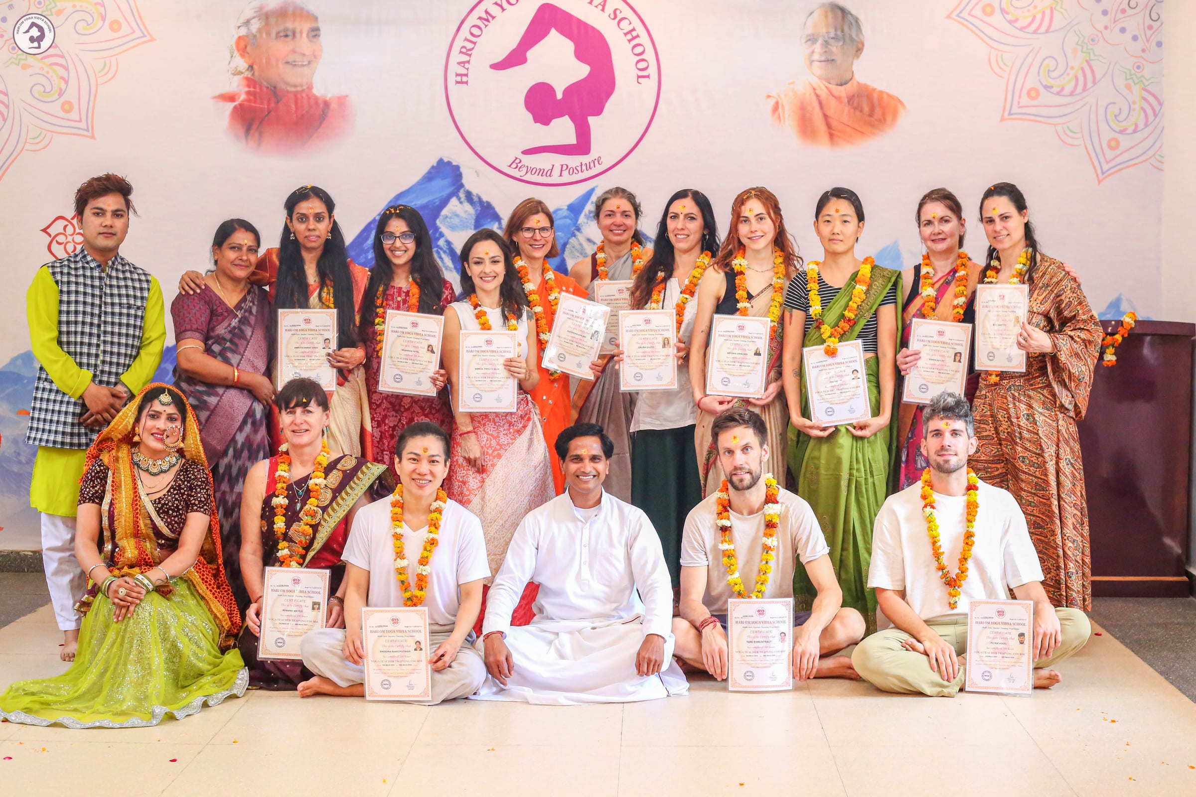 Graduates of a Hariom Yoga Vidya School teacher training holding their certificates, with the founder seated in the middle of the front row, under the school's banner in Rishikesh