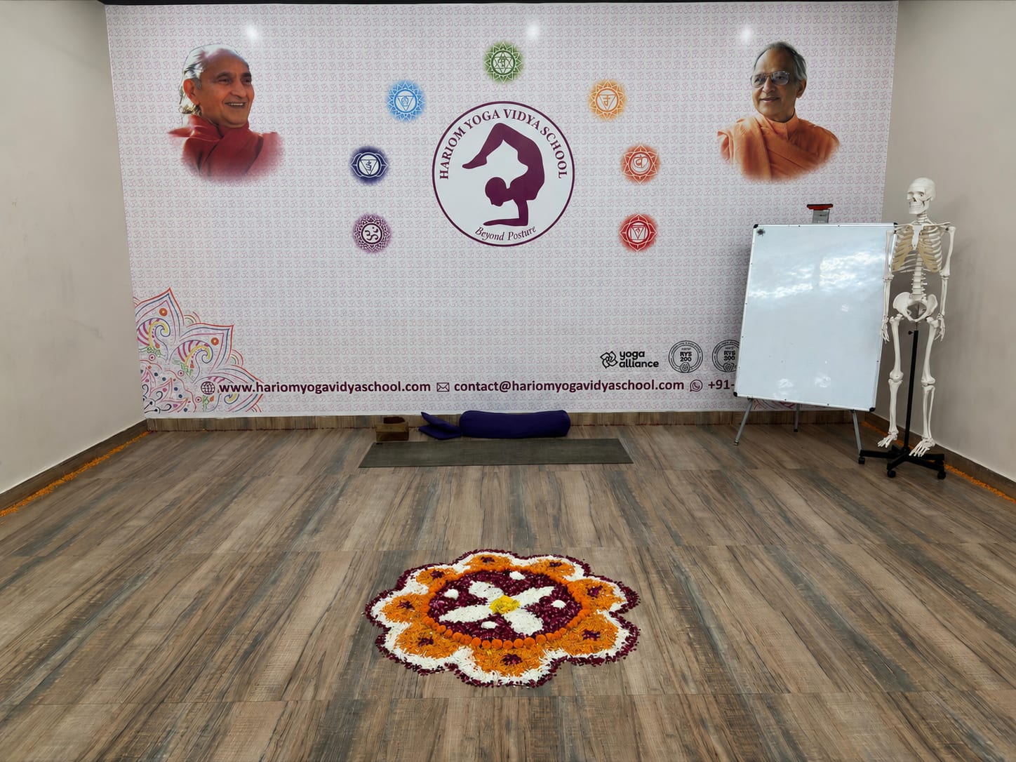 The yoga hall at Hariom Yoga Vidya School, Rishikesh: the school emblem and the founders' portraits on the welcome wall, a flower rangoli laid out on the wooden floor